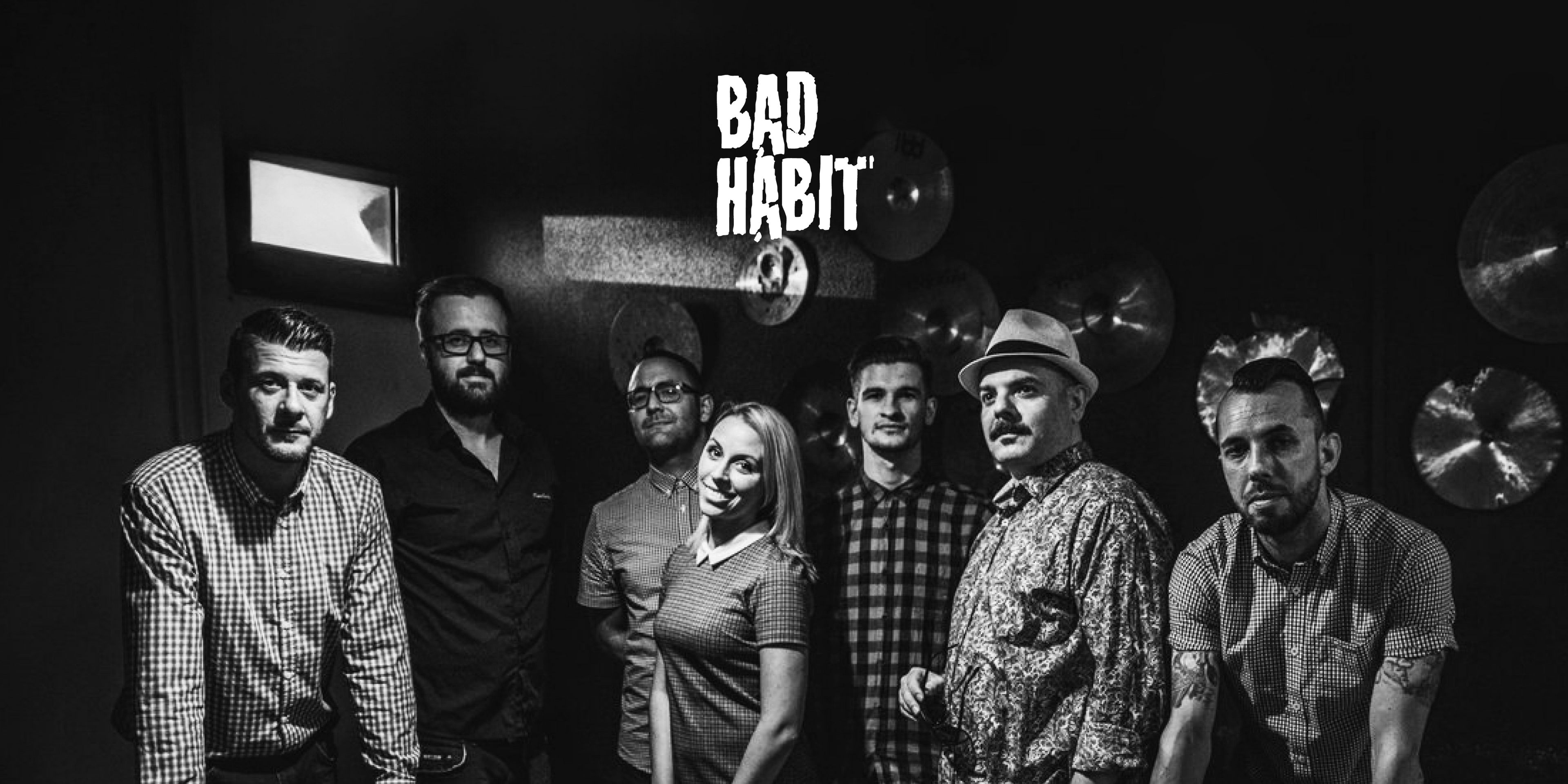 Die Band Bad Habit. Sieben Personen mit Hemden stehen an einem Billard-Tisch, im Hintergrund hängen Becken an der Wand, oben in der Mitte das Logo der Band.