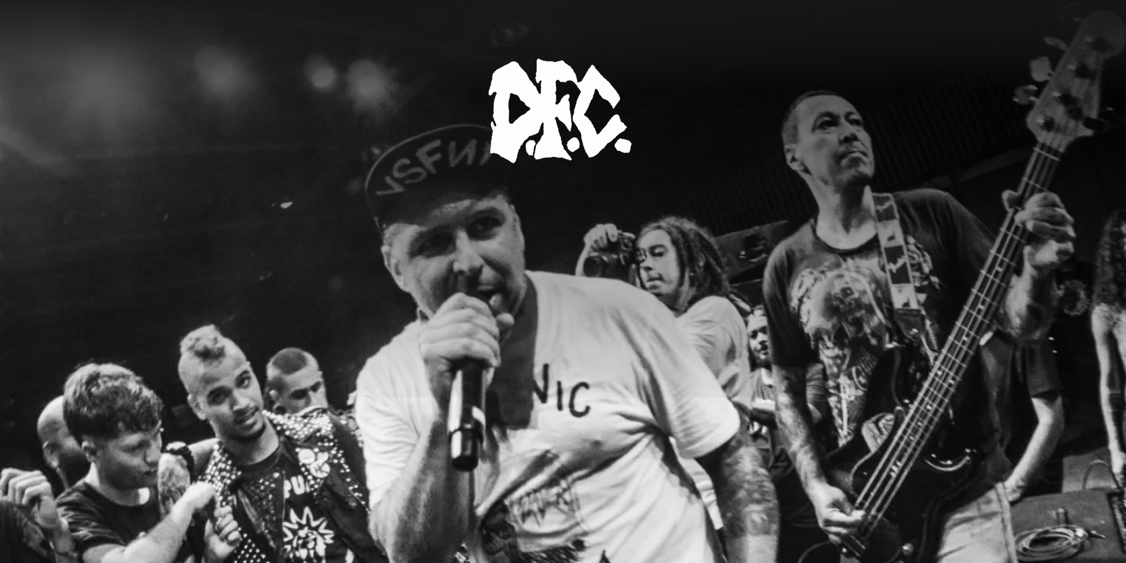 Die Band "DFC" während einem Live-Auftritt, eine Person hält ein Mikrofon, eine Person einen Bass, im Hintergrund Publikum. Oben in der Mitte das Logo der Band.