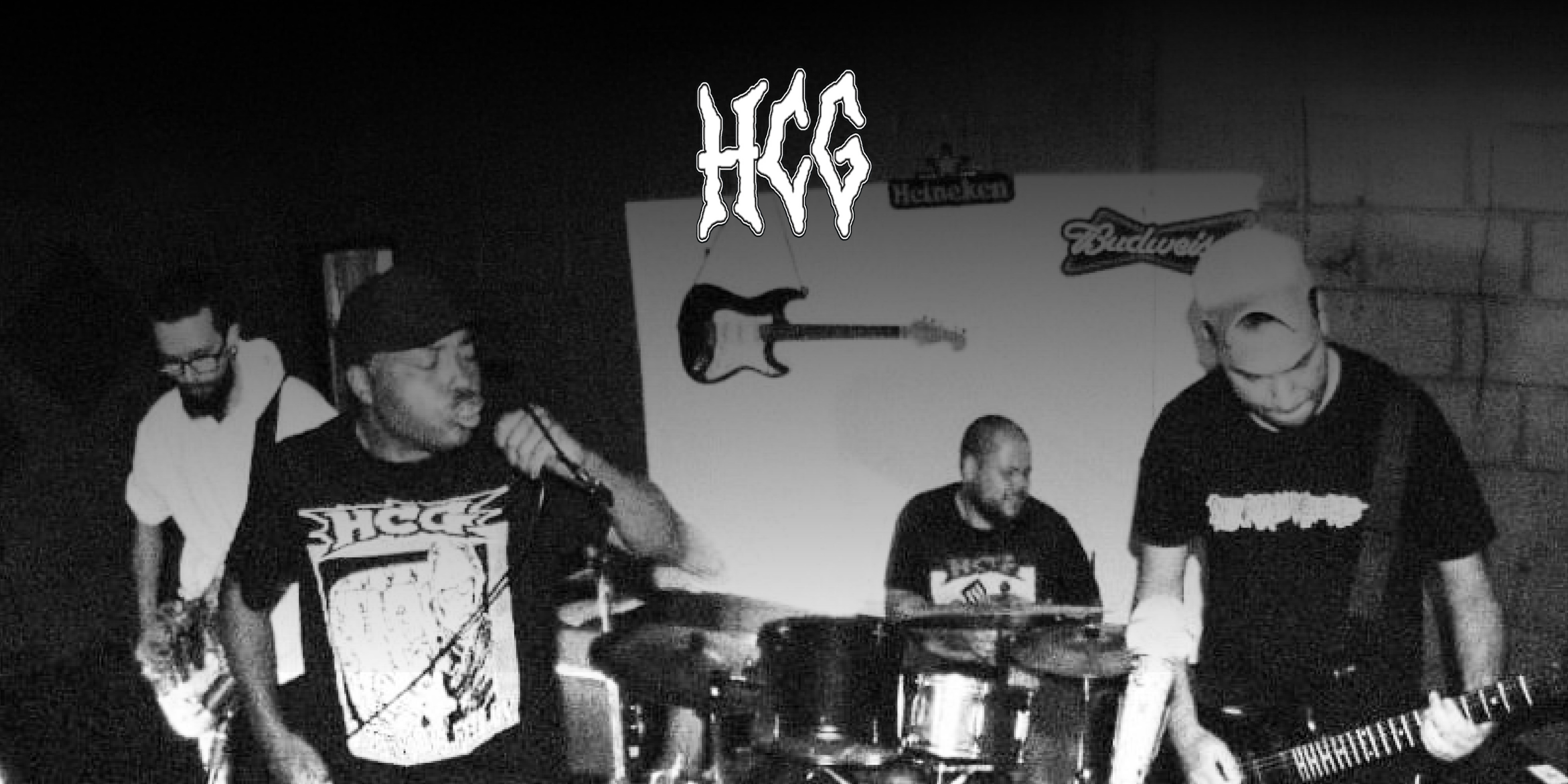 Die Band "HCG". Vier Personen während sie musizieren, links im Hintergrund eine Person am Bass, rechts davon im Vordergrund eine Person mit Mikrofon. Im Zentrum hinten eine Person am Schlagzeug und rechts im Vordergrund eine Person mit Gitarre. Oben in der Mitte das Logo der Band.