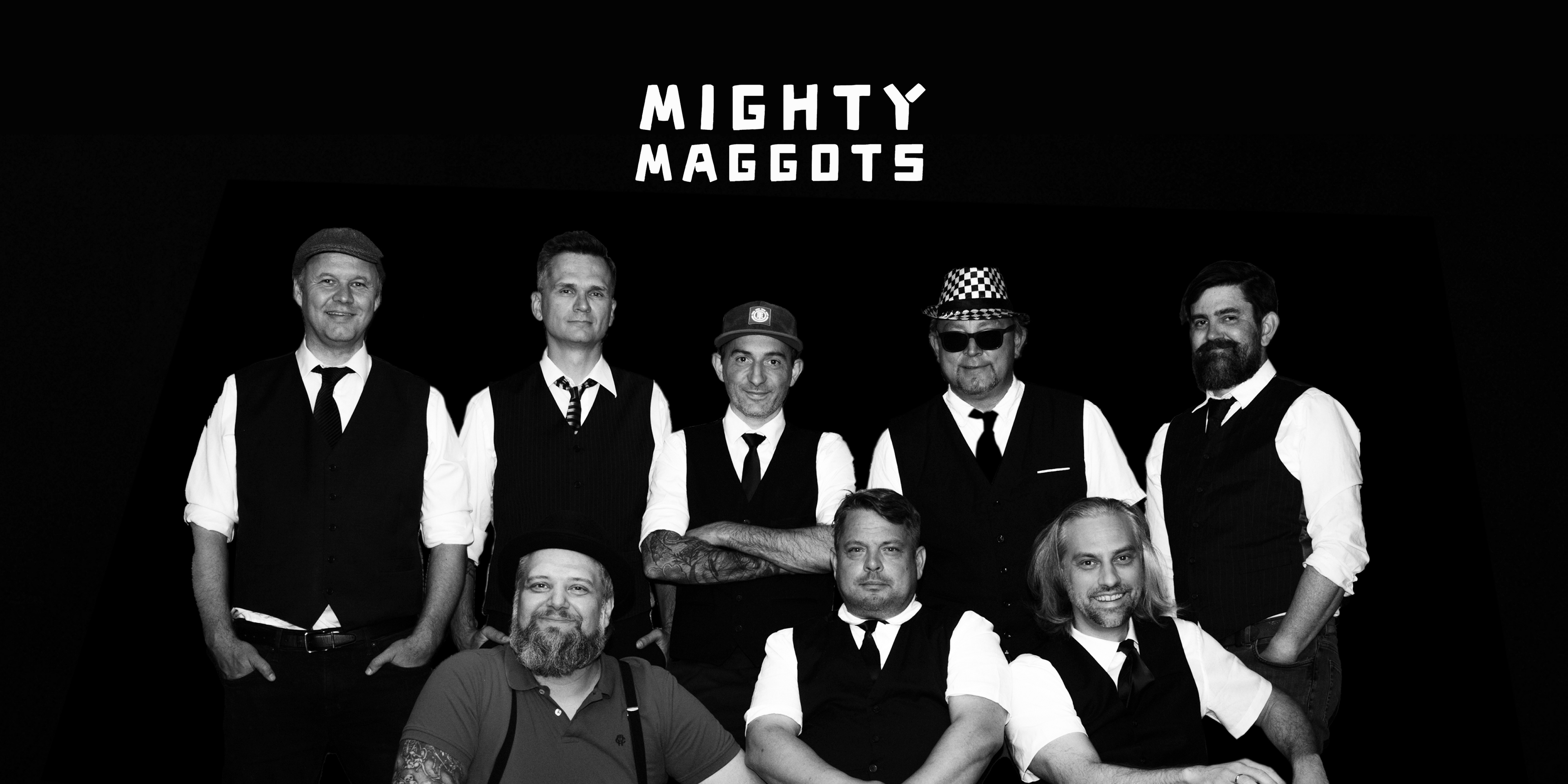 Die Band "Mighty Maggots". Acht Personen (fünf im Hintergrund, drei im Vordergrund) mit Hemden, Gilets und Krawatten blicken in die Kamera. Oben in der Mitte das Logo der Band.