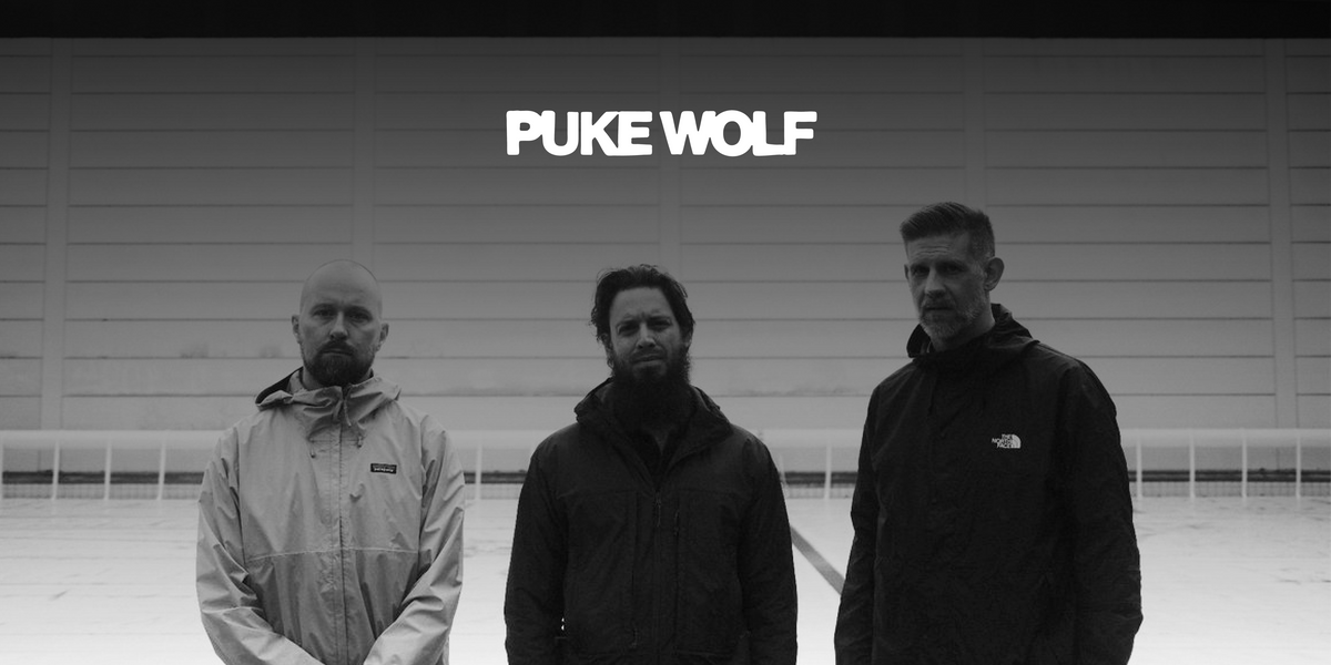 Die Band "Puke Wolf". Drei Männer stehen nebeneinander vor einem weißen Gebäude, blicken in die Kamera. Darüber in weiß das Logo der Band.